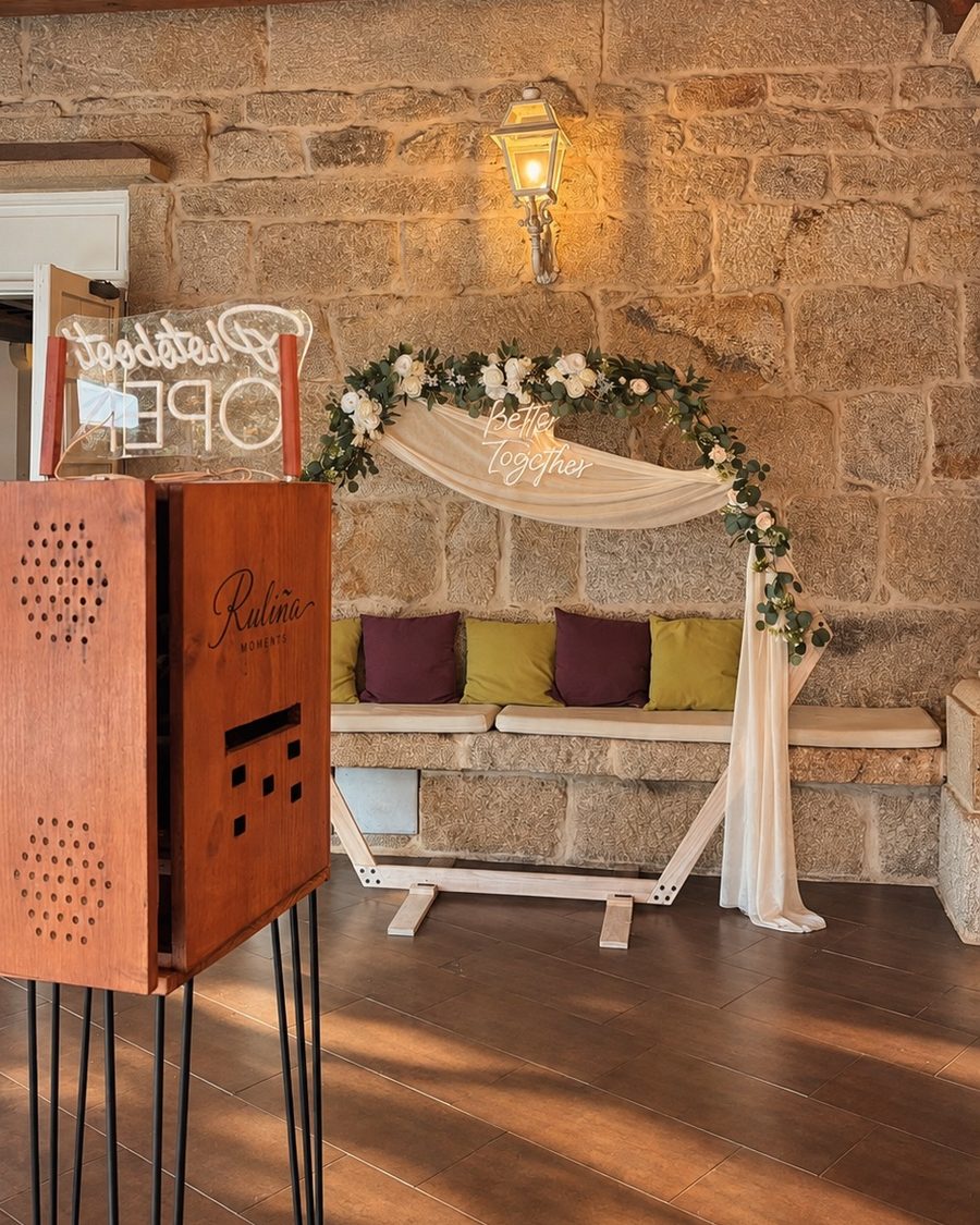 Fotomatón de madera de Ruliña Moments junto a un arco decorativo con flores y neón Better Together en un salón de piedra